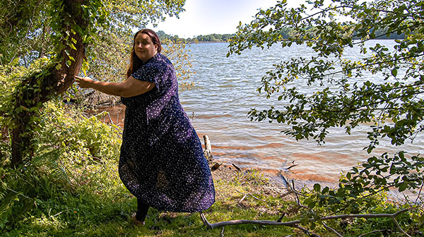 Photo de Pauline, magnétiseuse, debout près d'un arbre au bord d'un lac. Elle semble en pleine interaction avec la nature, dans un cadre paisible et lumineux, évoquant une connexion spirituelle et énergétique avec les éléments environnants.