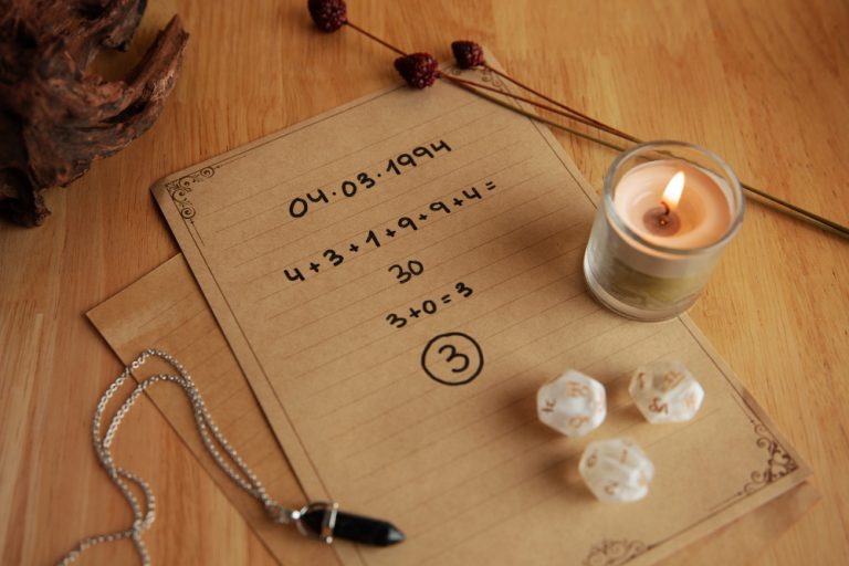 Photo illustrant une séance de numérologie avec une date de naissance écrite sur du papier, des calculs réalisés pour trouver un chiffre personnel, accompagnée d'une bougie allumée, d'un pendule et de dés mystiques. Une ambiance chaleureuse et spirituelle se dégage de la composition.