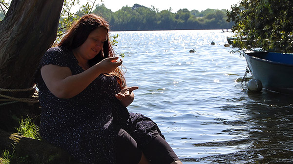 Photo de Pauline, magnétiseuse, assise au bord d'un lac avec un pendule dans la main. Elle semble en pleine pratique énergétique, utilisant le pendule pour une séance de radiesthésie ou d'harmonisation, dans un cadre naturel apaisant.